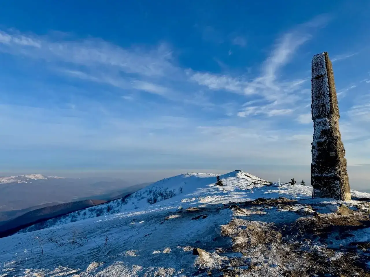 Kamienny obelisk na szczycie ośnieżonych **Rawki Bieszczady** pod błękitnym niebem.