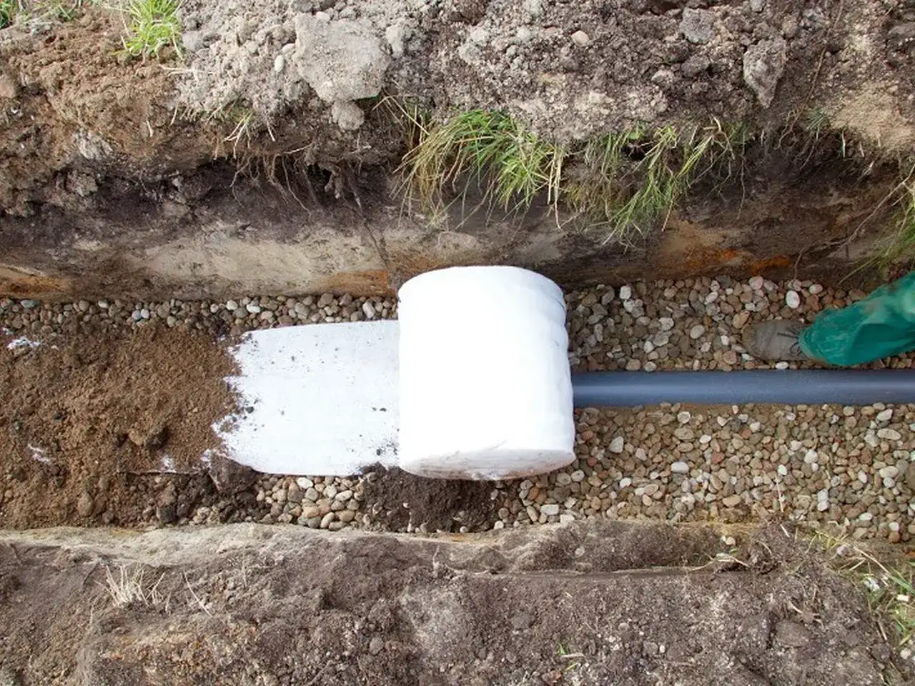 Budowa drenażu: rura drenarska owinięta geowłókniną, otoczona żwirem i ziemią, gotowa do zasypania.