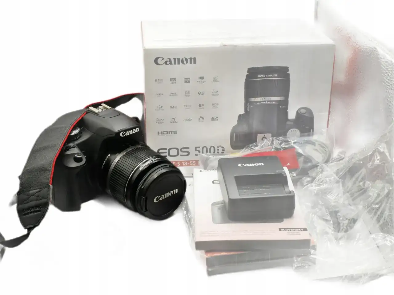 Aparat Canon EOS 500D z obiektywem i akcesoriami na białym tle.