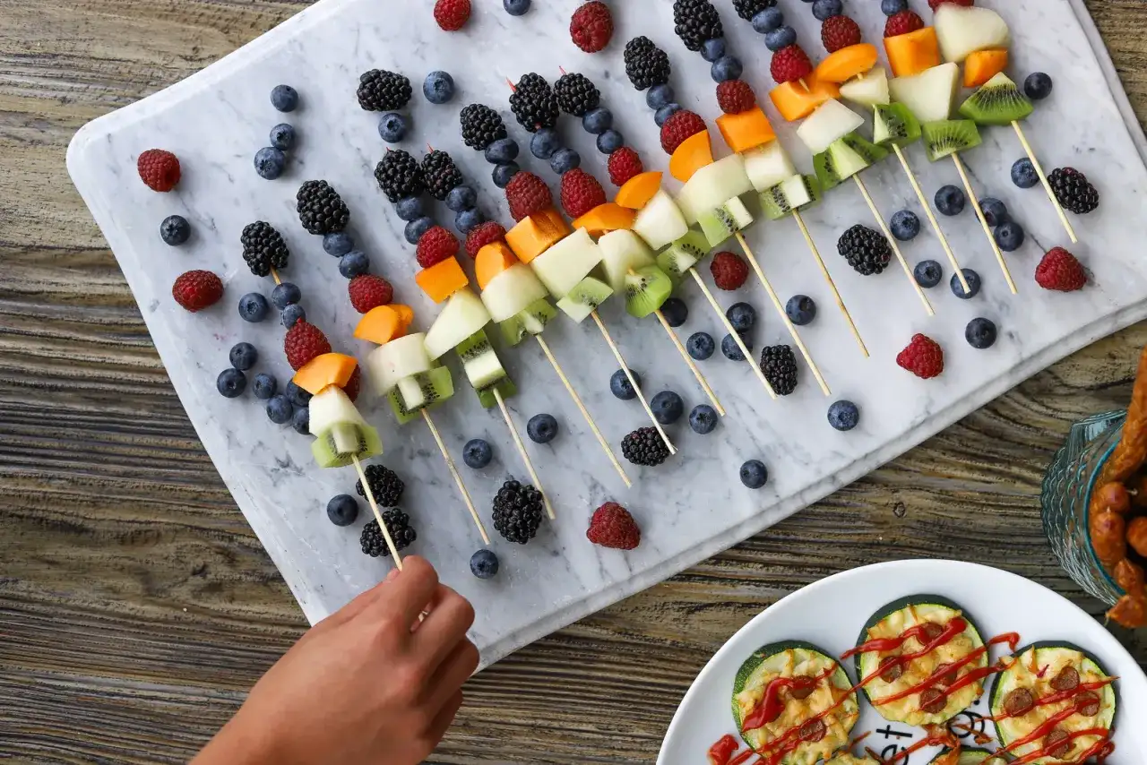 Kolorowe szaszłyki owocowe z kiwi, melonem, malinami, borówkami i jeżynami na marmurowej desce.