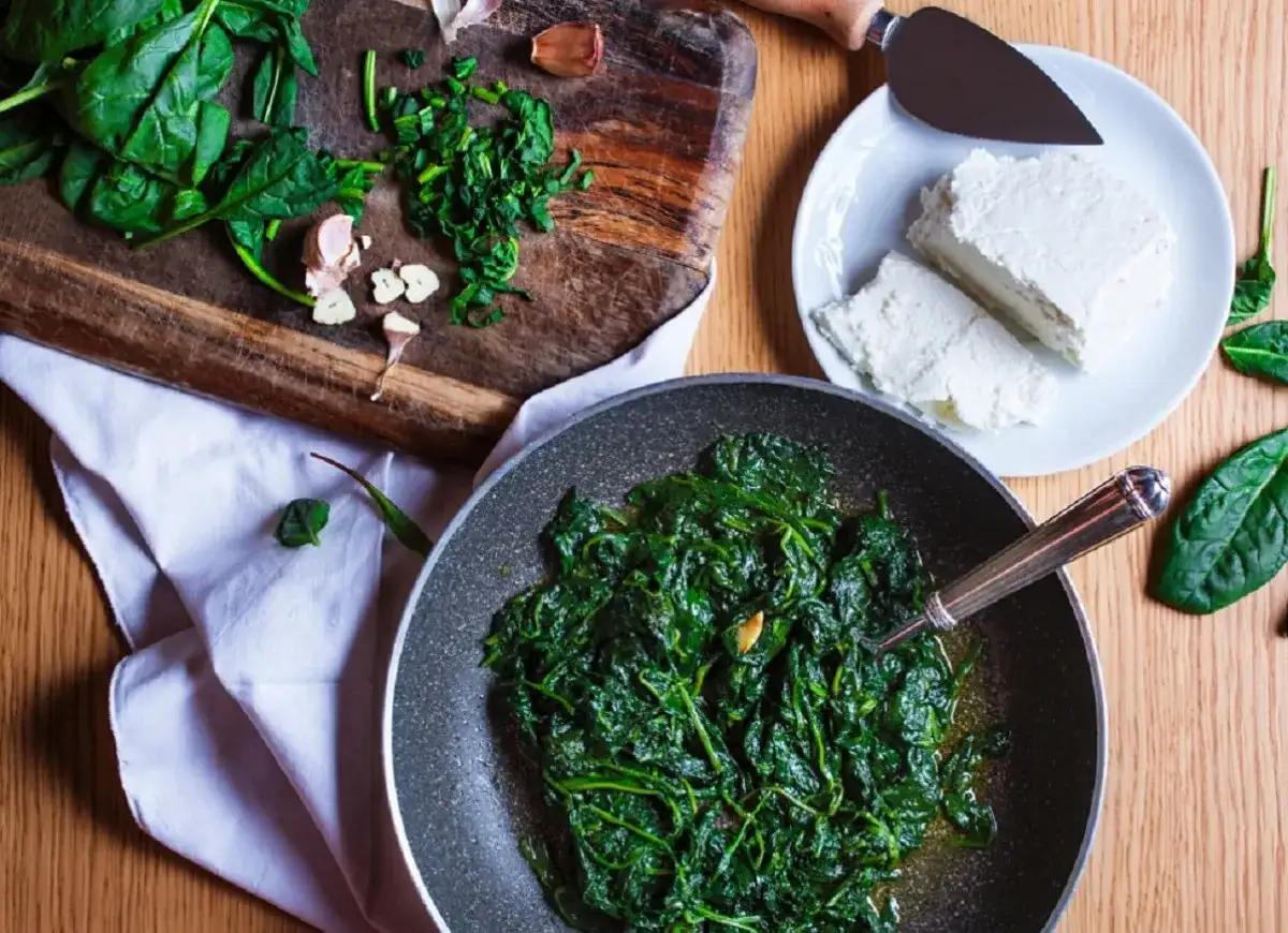 Jak doprawić szpinak? Na desce świeży szpinak, czosnek i ser feta na talerzu. W patelni gotowy, duszony szpinak.