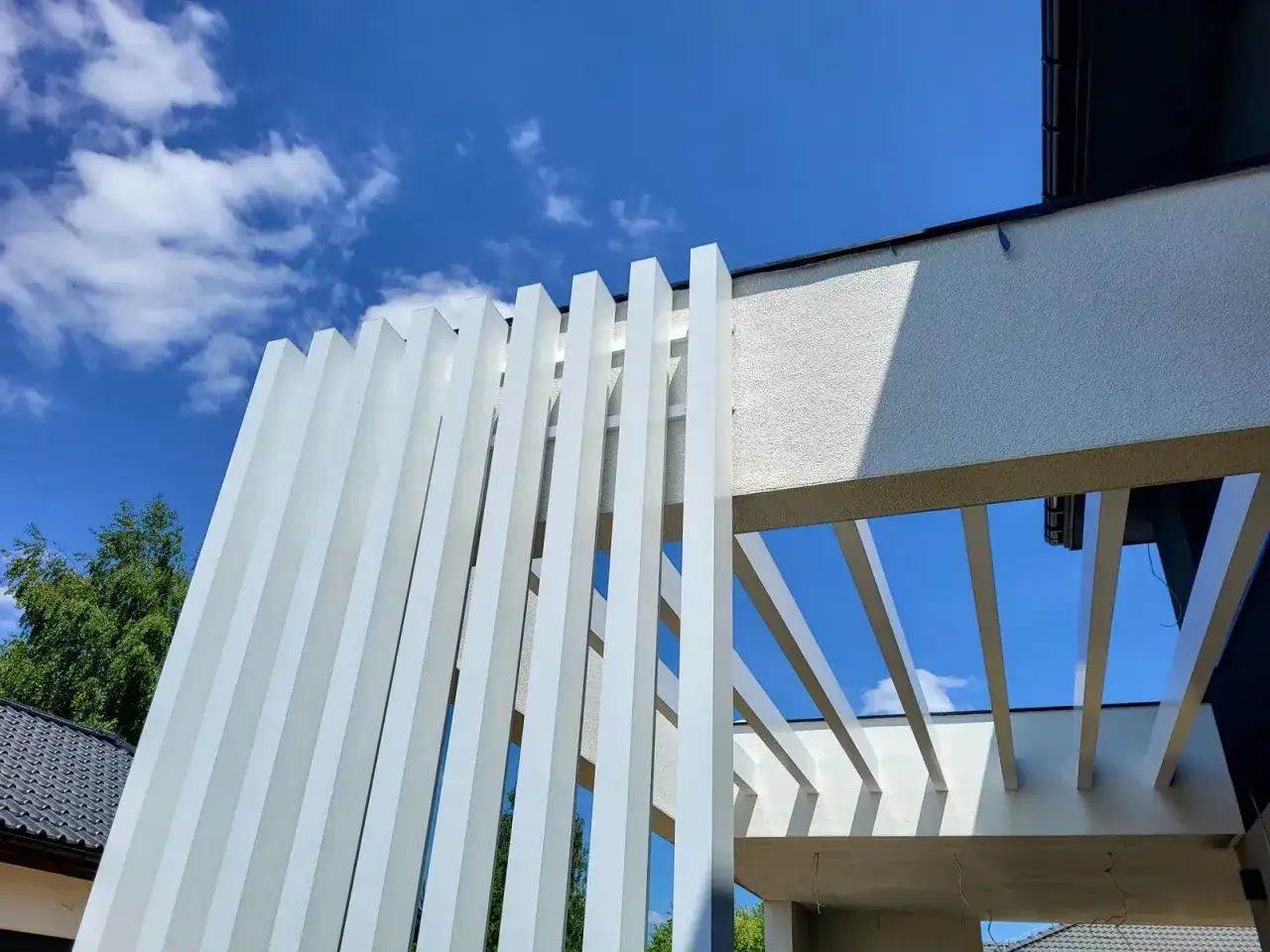 Nowoczesna pergola z białymi, pionowymi lamelami, wykonana z **belki aluminiowe**. Konstrukcja tworzy ciekawy wzór na tle błękitnego nieba z chmurami.