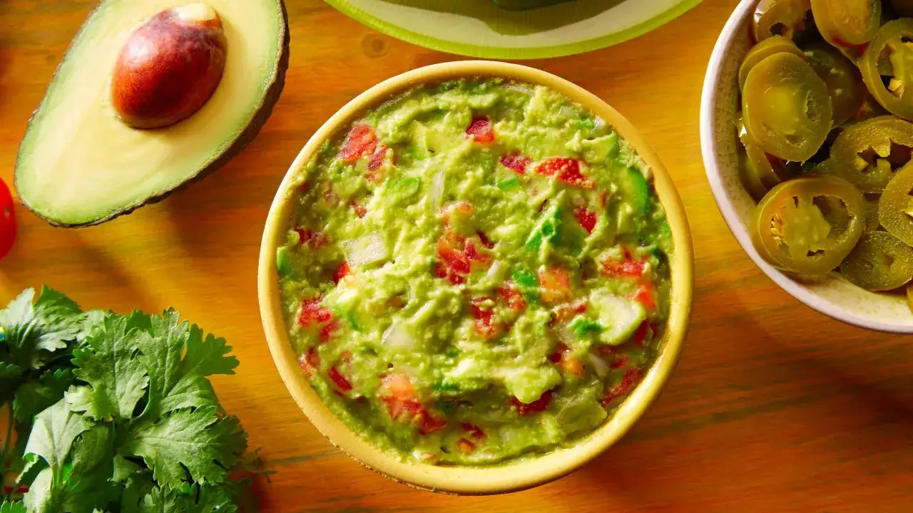 Miseczka guacamole z awokado, pomidorami i cebulą, obok połówka awokado i plasterki jalapeño.