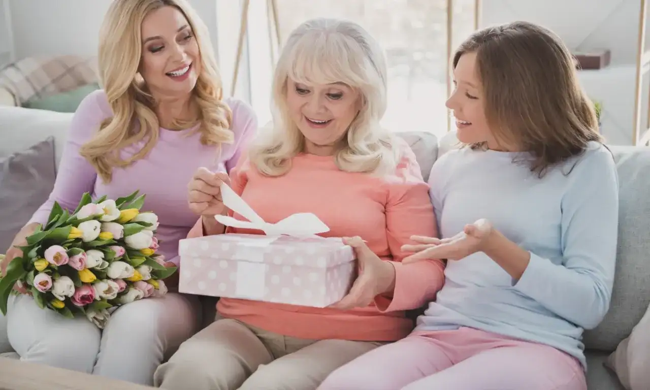 Trzy kobiety na kanapie: starsza otwiera prezent, młodsza trzyma bukiet, najmłodsza gestykuluje.