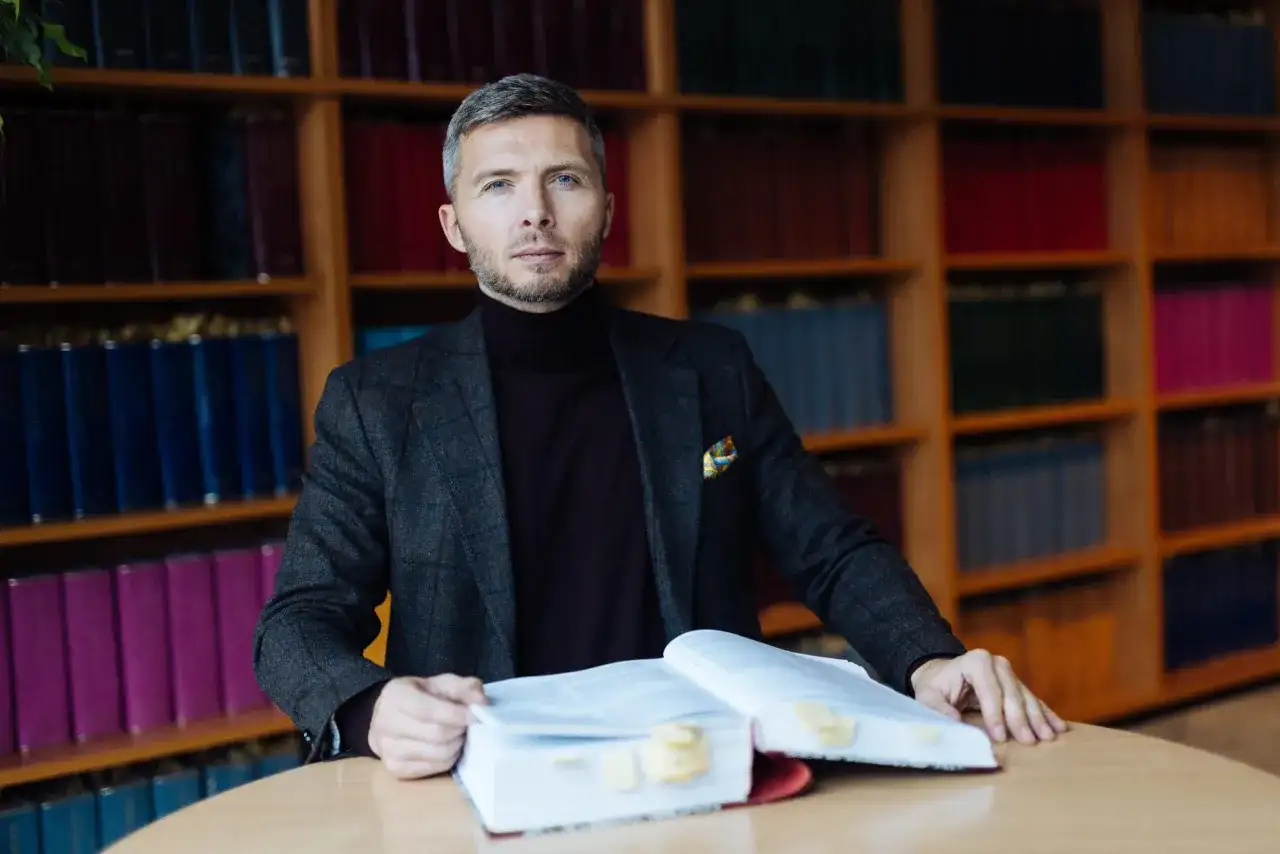 Mężczyzna w garniturze siedzi przy stole z otwartą księgą, otoczony regałami pełnymi książek. To jego zabezpieczenie STB.