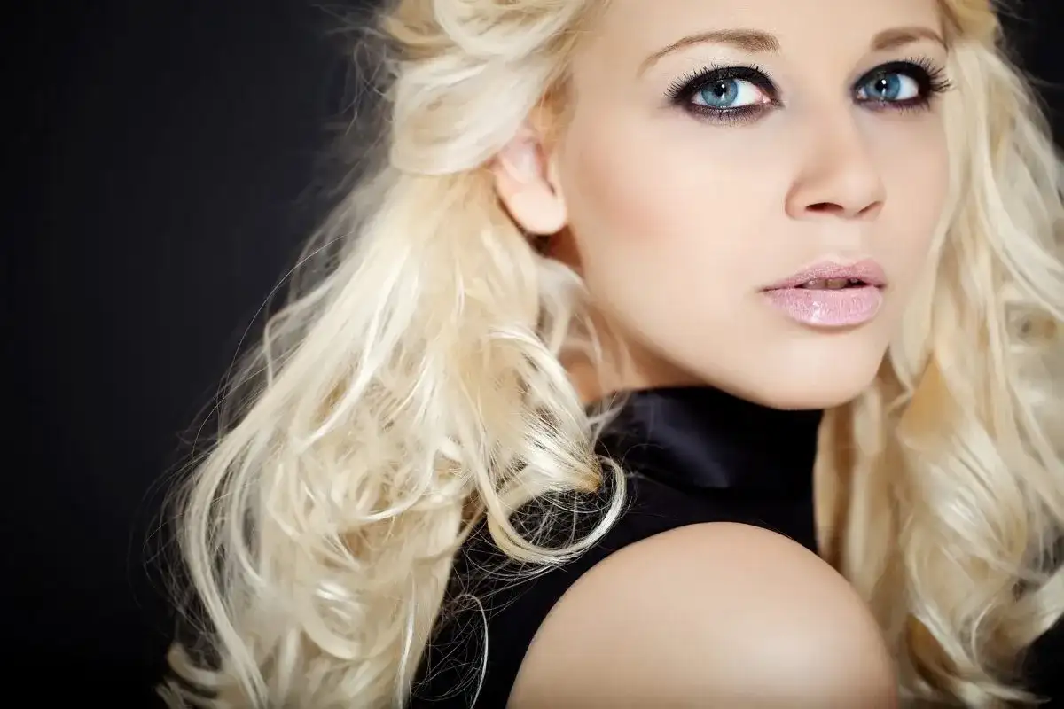 Blondynka z niebieskimi oczami i ciemną oprawą oczu, w czarnej sukience, na czarnym tle.