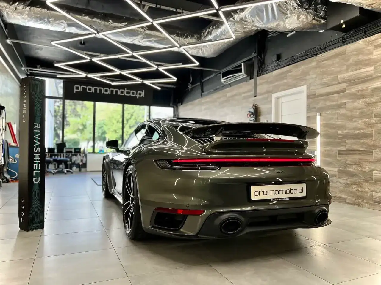 Ciemnozielone Porsche 911 w garażu z nowoczesnym oświetleniem i stojącym obok pionowym banerem.