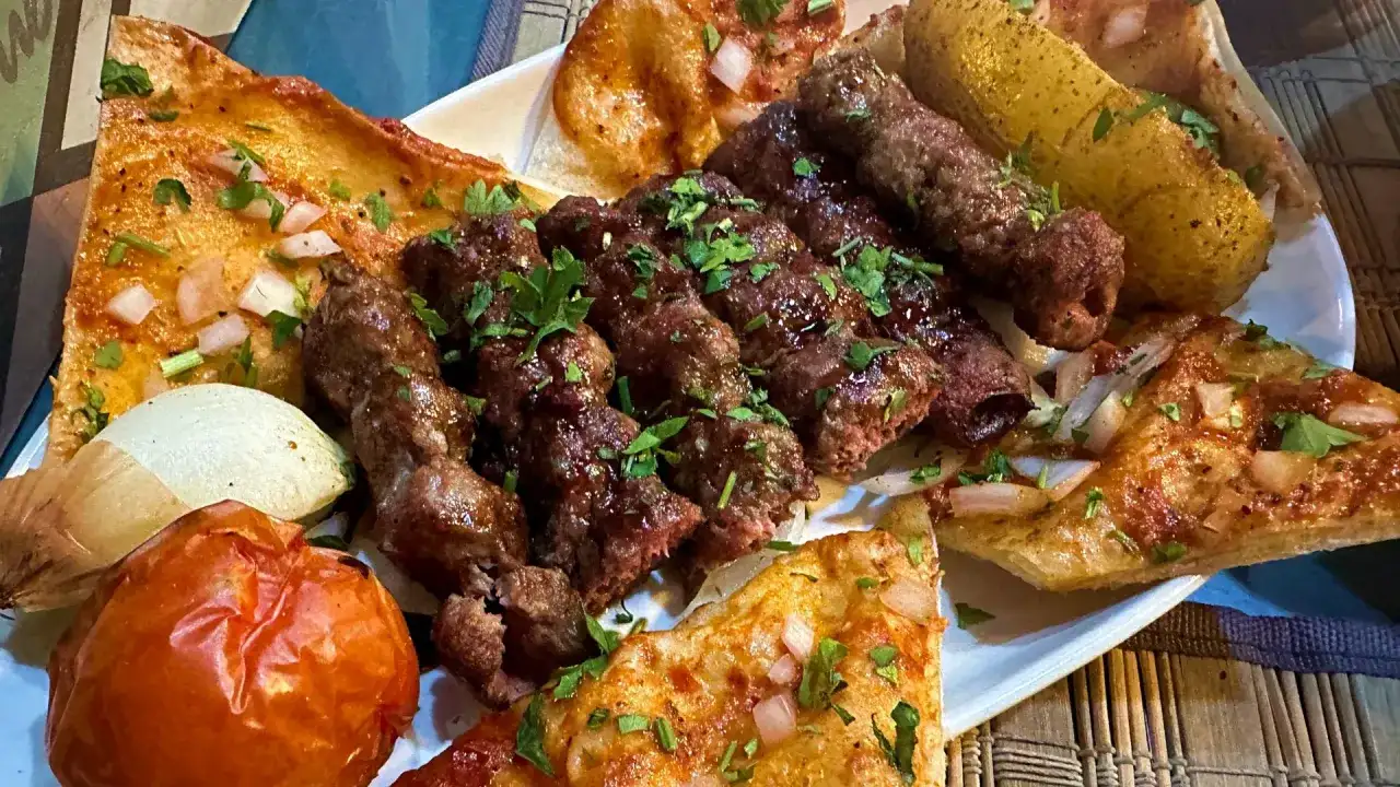 Talerz z grillowanymi szaszłykami jagnięciny, pieczonymi ziemniakami, pomidorem i chlebem.