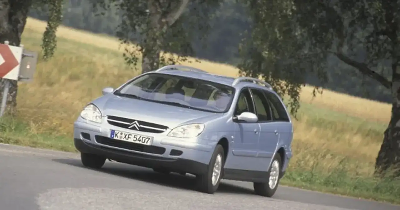 Jasnoniebieskie kombi Citroen na zakręcie drogi, z drzewami i polem w tle.