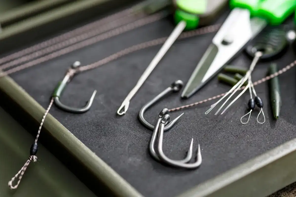 Zbliżenie na haczyki karpiowe, żyłkę, nożyczki i inne akcesoria wędkarskie w pudełku.