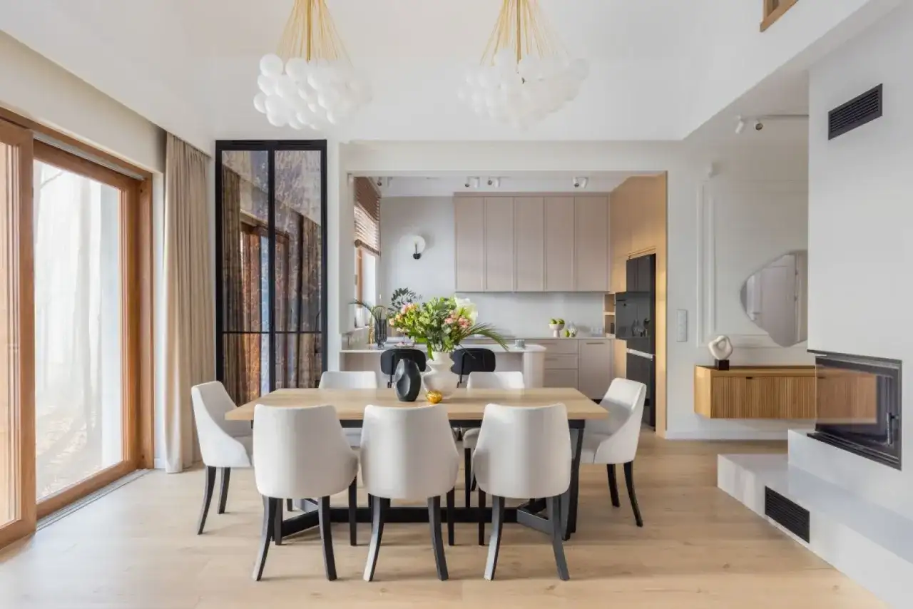 Nowoczesne oświetlenie salonu z jadalnią aranżacje: drewniany stół, białe krzesła, kuchnia w tle, kominek.