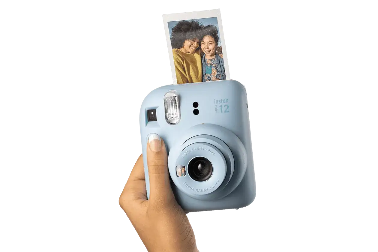 Niebieski aparat Instax Mini 12, trzymany w dłoni, z właśnie wysuniętym, jasnym zdjęciem dwóch uśmiechniętych przyjaciółek.