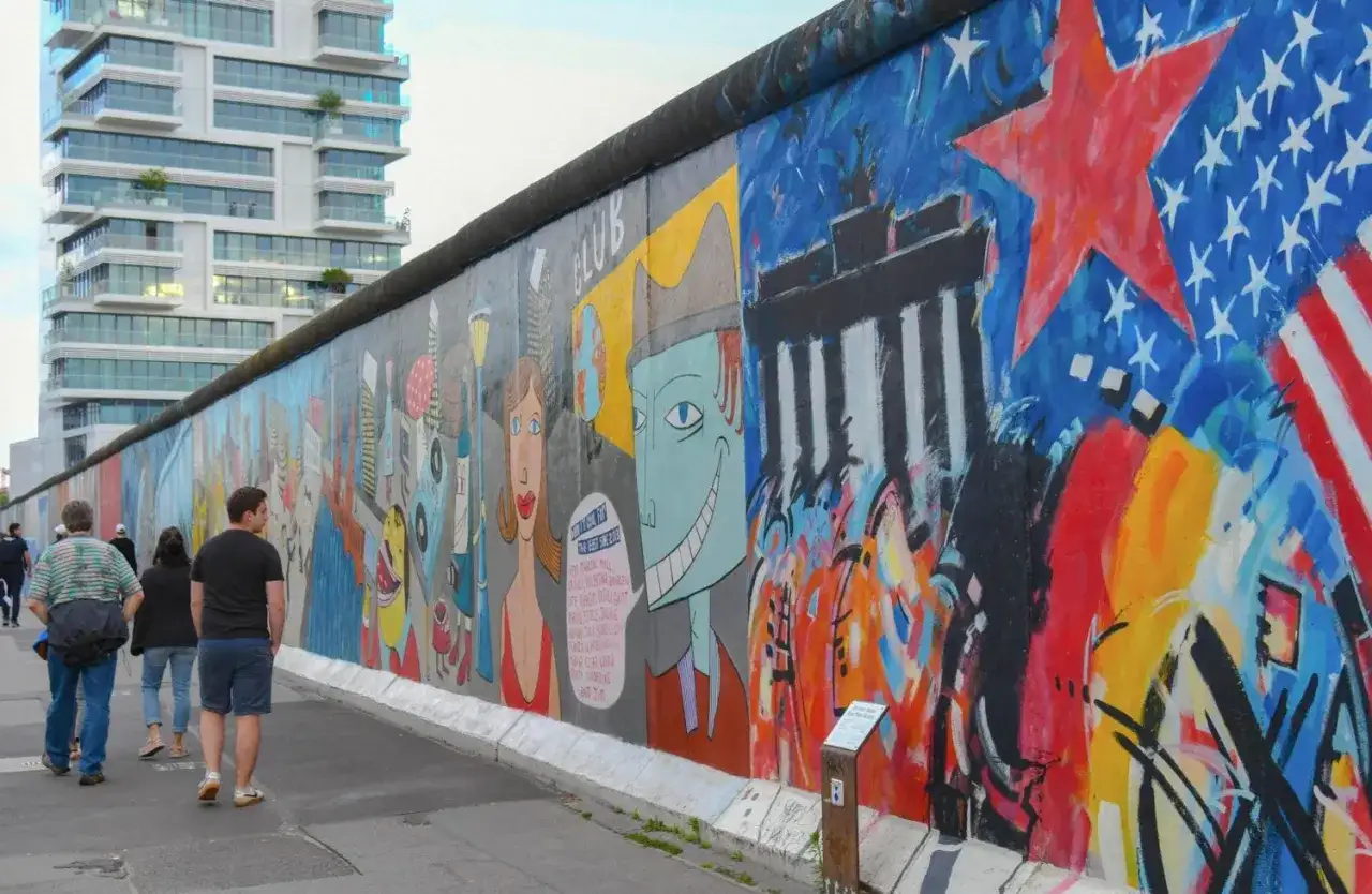 Bunte East Side Gallery in Berlin mit Graffiti und Menschen, im Hintergrund ein modernes Hochhaus.