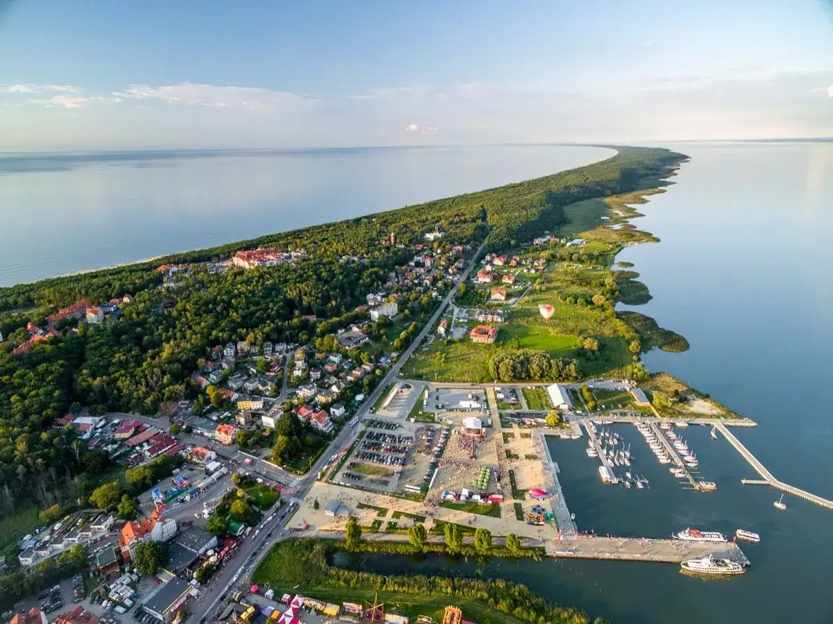 Widok z lotu ptaka na Krynicę Morską. Widać zalew, lasy, zabudowania i tętniący życiem rynek z mariną pełną łodzi.