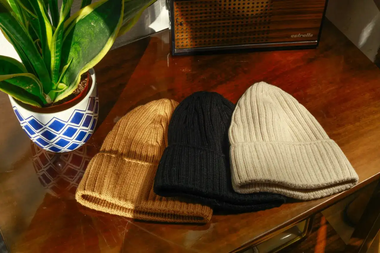 Trzy ciepłe czapki z merynosa w kolorach karmelowym, czarnym i ecru leżą obok siebie na drewnianym stole.