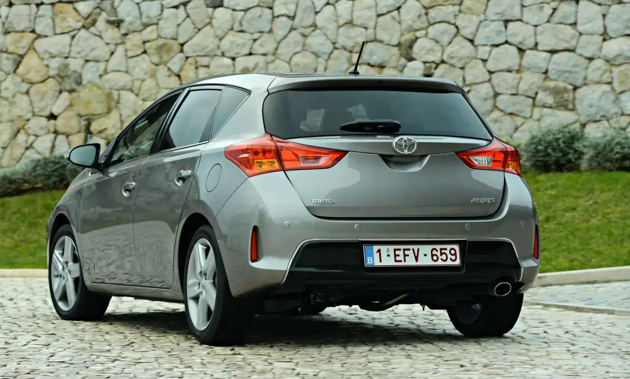 Tył szarego samochodu Toyota Auris z belgijską tablicą rejestracyjną na tle kamiennej ściany.