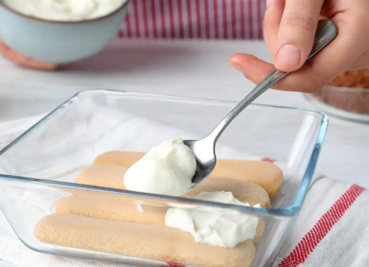 Nakładanie łyżką puszystego kremu z mleka skondensowanego i mascarpone na biszkopty w szklanej formie.