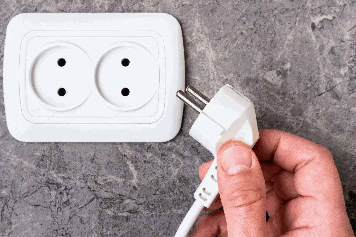 Dłoń trzyma wtyczkę do podłączenia pralki. Gniazdko elektryczne znajduje się na ścianie.