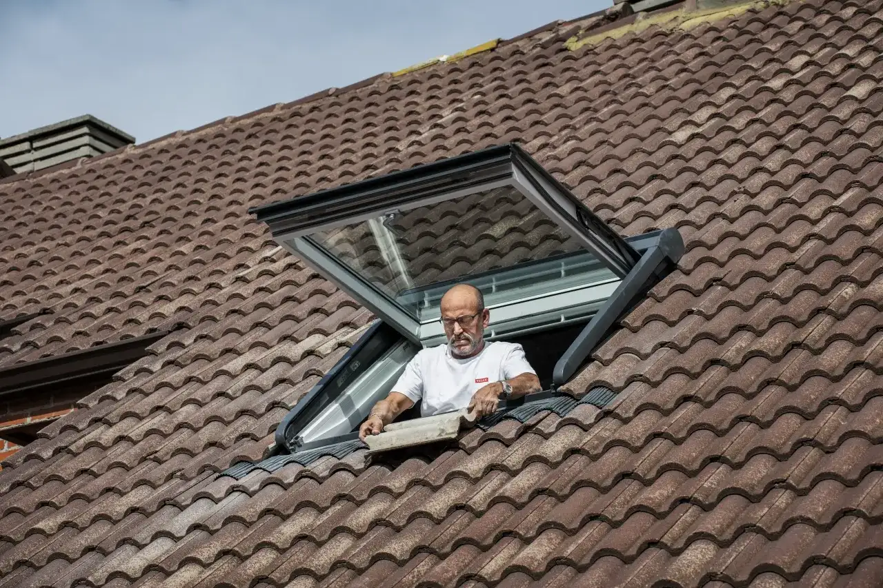 Mężczyzna montuje okno dachowe balkonowe na brązowym, dachówkowym dachu.