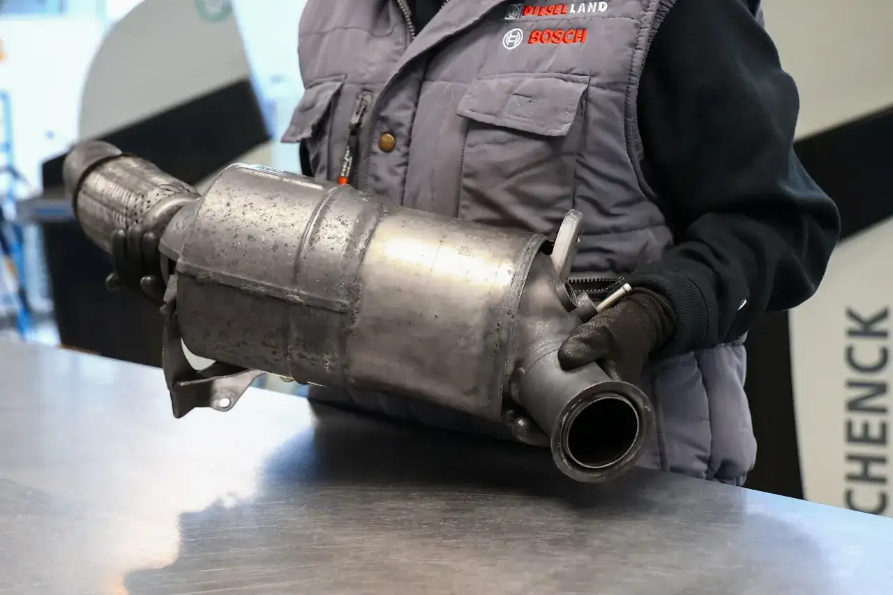 Mechanik w rękawiczkach trzyma metalowy element układu wydechowego samochodu.