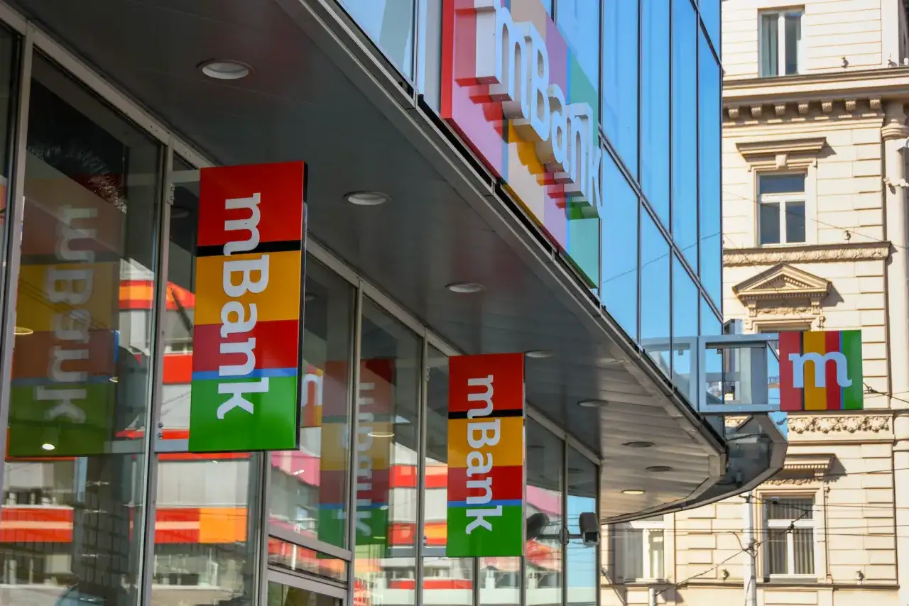 Baner mBank z kolorowym logo i napisem "mBank" na tle nowoczesnego budynku.