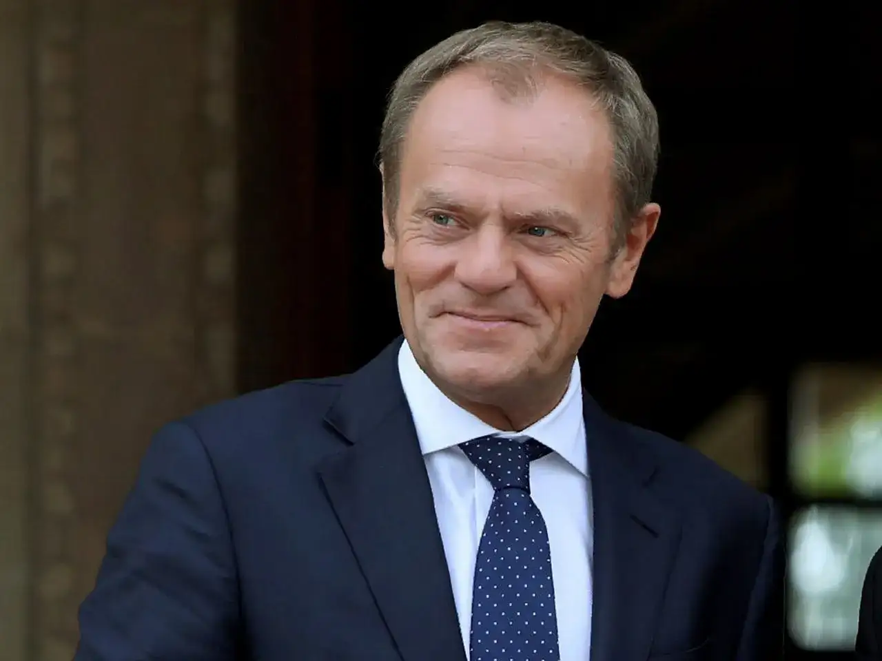 Uśmiechnięty Donald Tusk w granatowym garniturze i krawacie w kropki. Czy Tusk zna angielski? Pewnie tak, w końcu to polityk europejski!