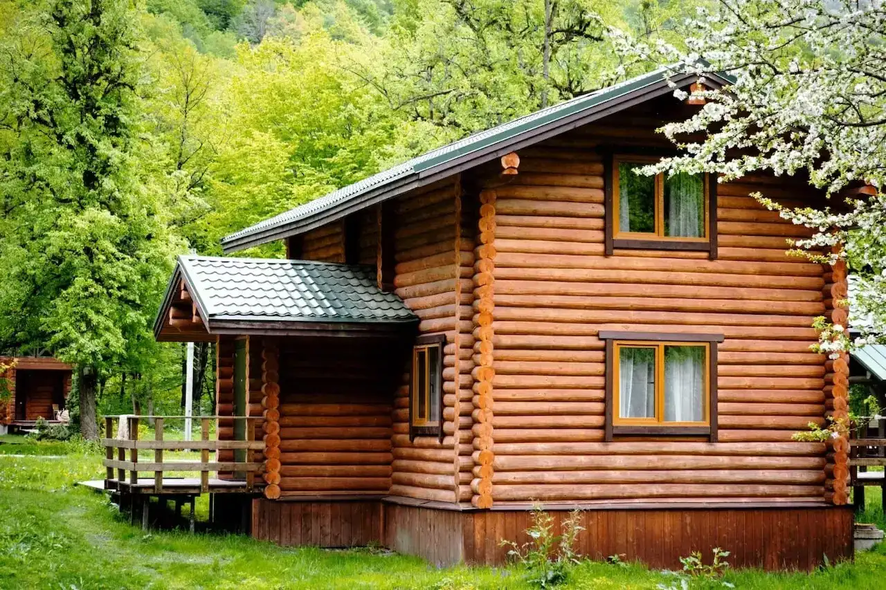 Drewniany dom letniskowy z zielonym dachem, otoczony zielenią i kwitnącym drzewem.