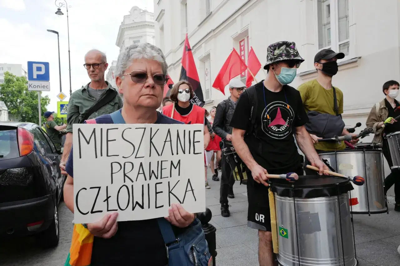 Kobieta trzyma transparent: "MIESZKANIE PRAWEM CZŁOWIEKA". W tle protestujący z bębnami i flagami.