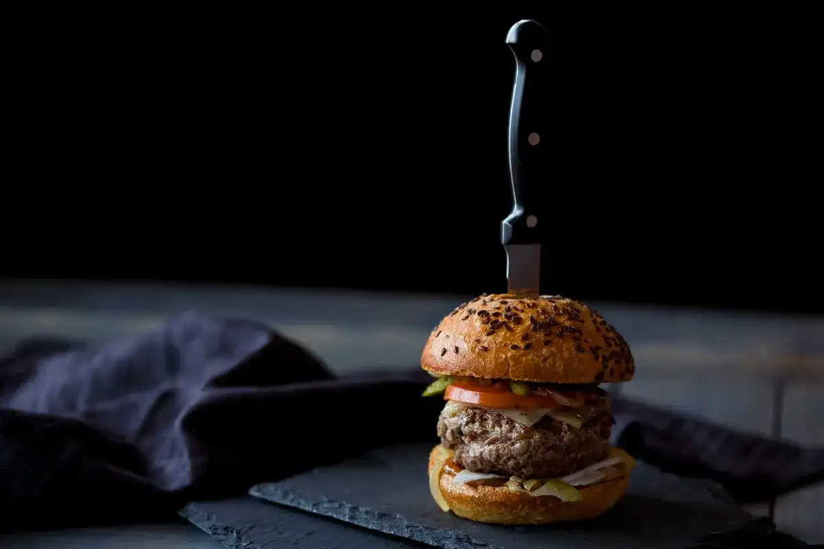 Soczysty burger wołowy z nożem wbitym w bułkę, na ciemnym tle.