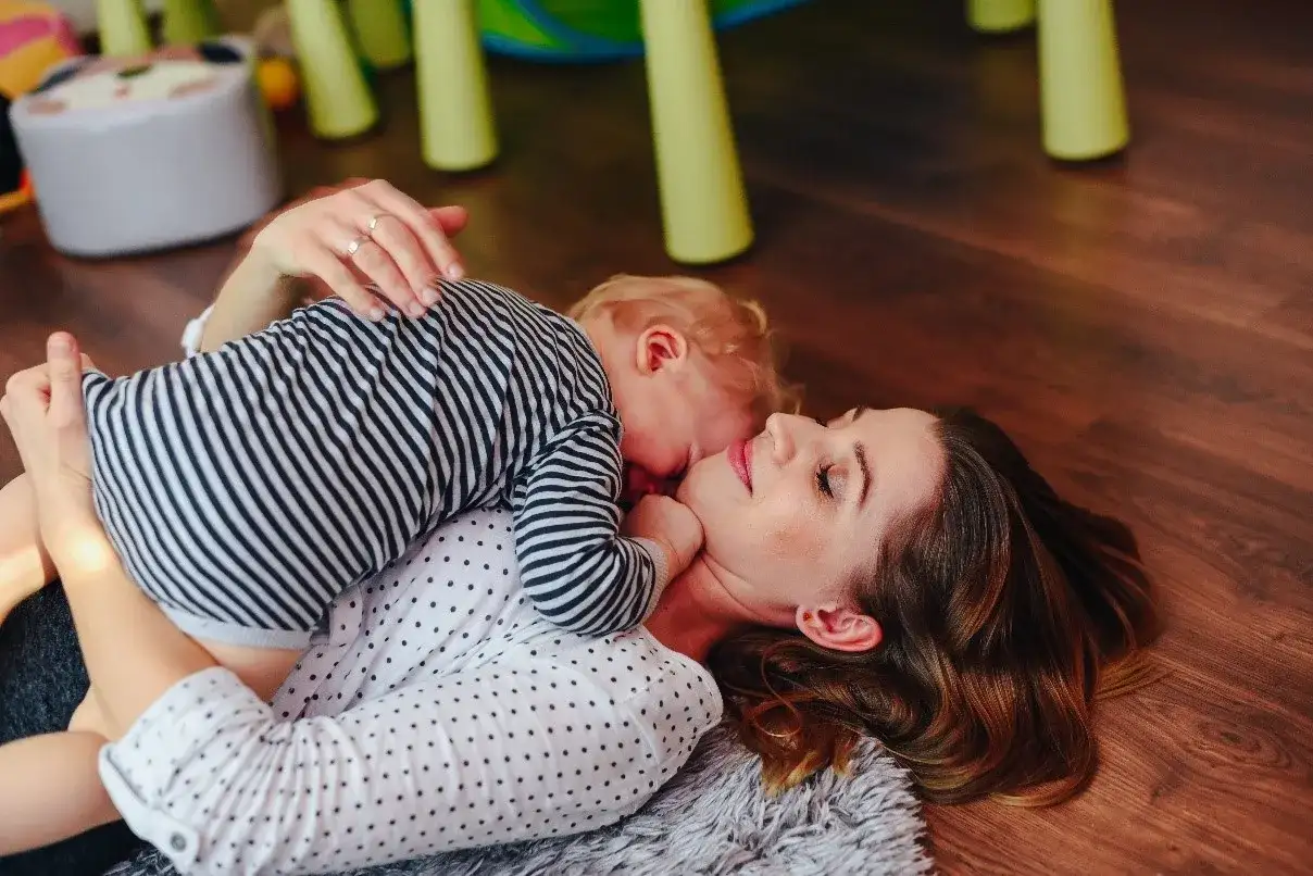 Mama i dziecko leżą na podłodze. Dziecko przytula się do mamy. To piękny przykład, co to stymulacja rozwoju poprzez bliskość i zabawę.