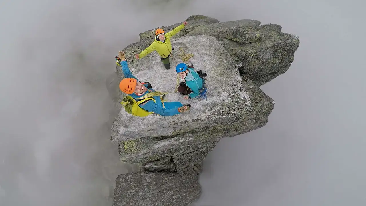 Trzech wspinaczy na skalnym występie, otoczonym mgłą. Jeden z nich, w niebieskim kombinezonie, wisi nad przepaścią, ciesząc się zdobyciem mnich szczyt.