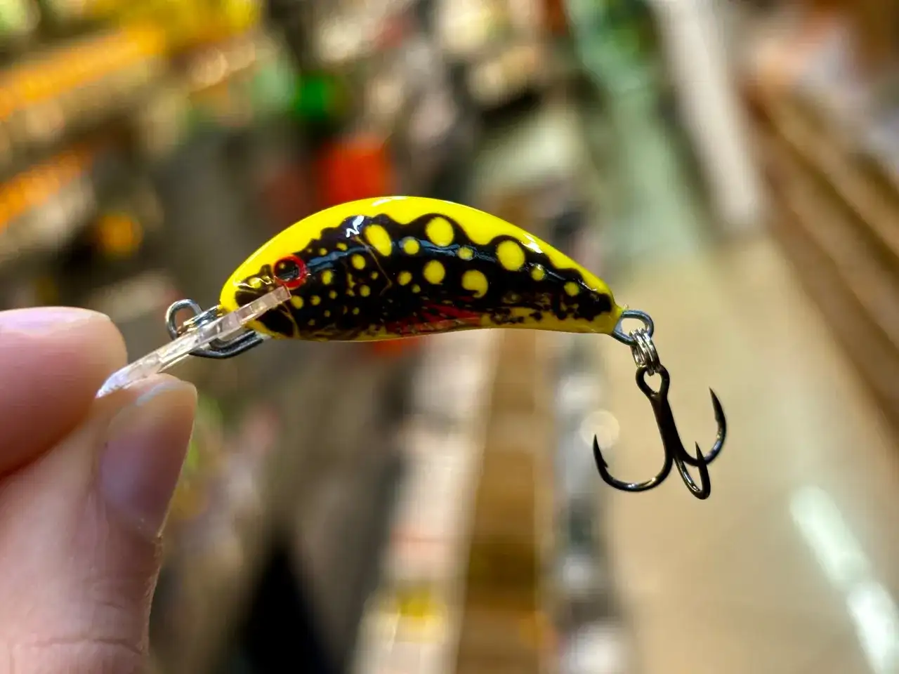 Żółta przynęta wędkarska z czarnymi plamkami i haczykiem, trzymana w palcach.