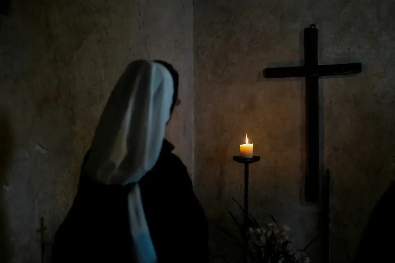 Zakonnica w ciemności przed krzyżem i płonącą świecą.