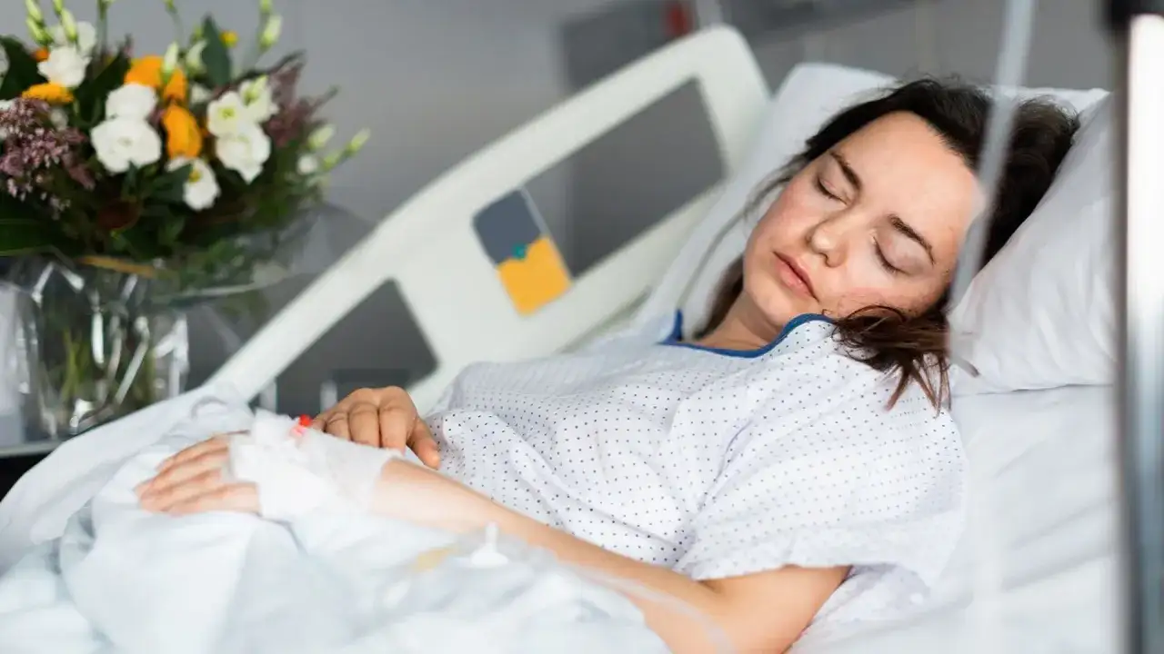 Kobieta w szpitalnym łóżku, z zamkniętymi oczami, ma założony wenflon. Obok bukiet kwiatów.