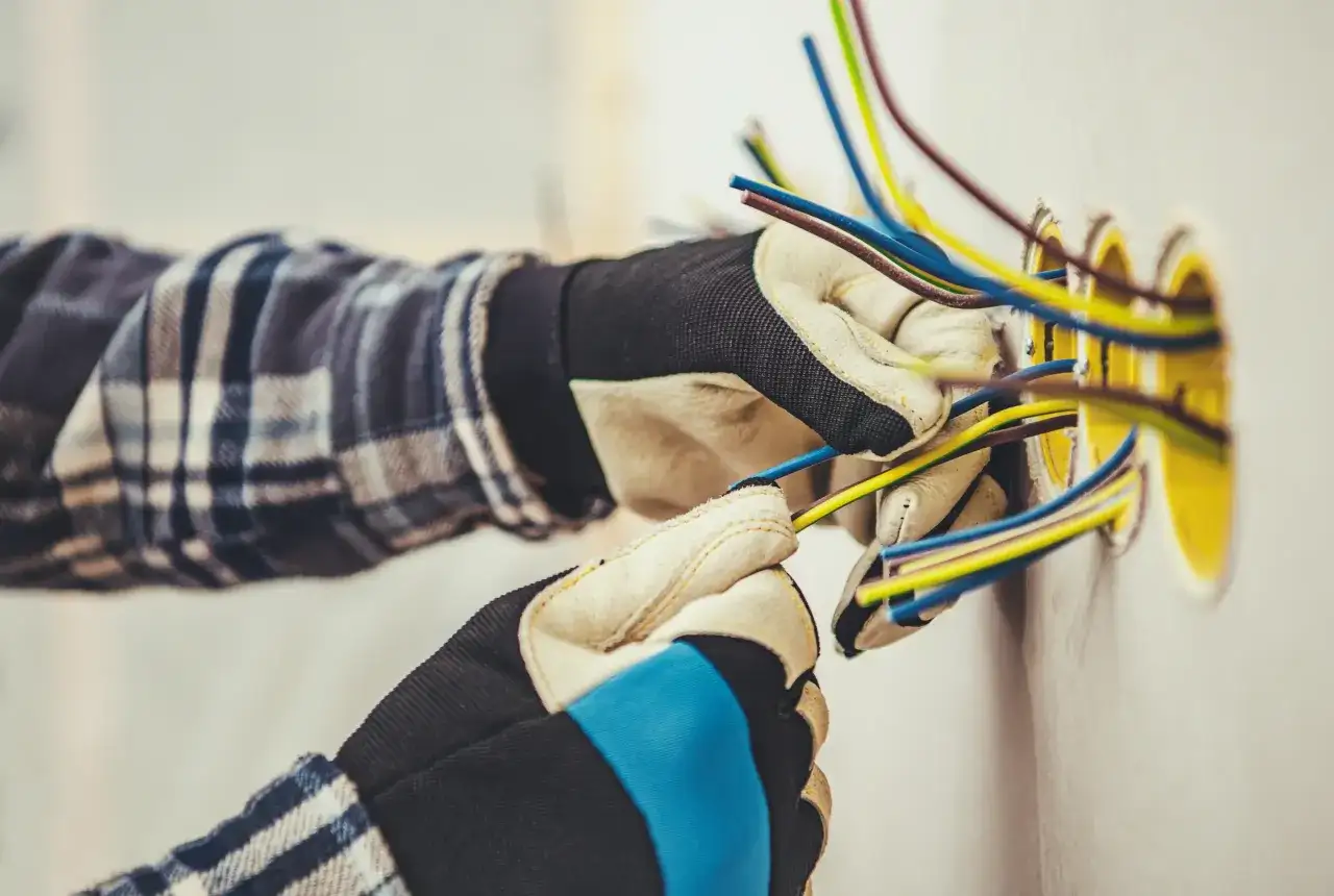 Elektryk w rękawicach przygotowuje przewody do podłączenia gniazdek. Widać kilka kolorowych kabli wychodzących ze ściany, gotowych do połączenia. To kluczowy etap, jak zaznaczyć gniazdka na projekcie.
