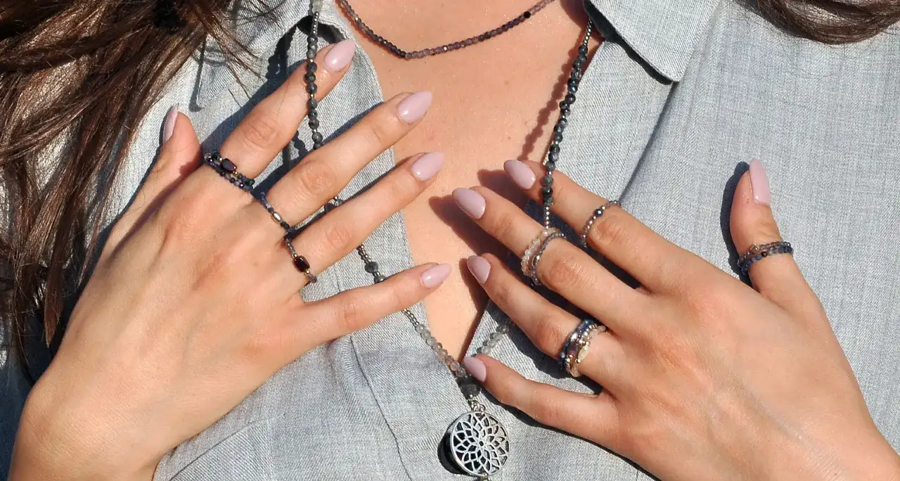 Dłonie z paznokciami w pastelowym różu, ozdobione pierścionkami i naszyjnikami z koralików.