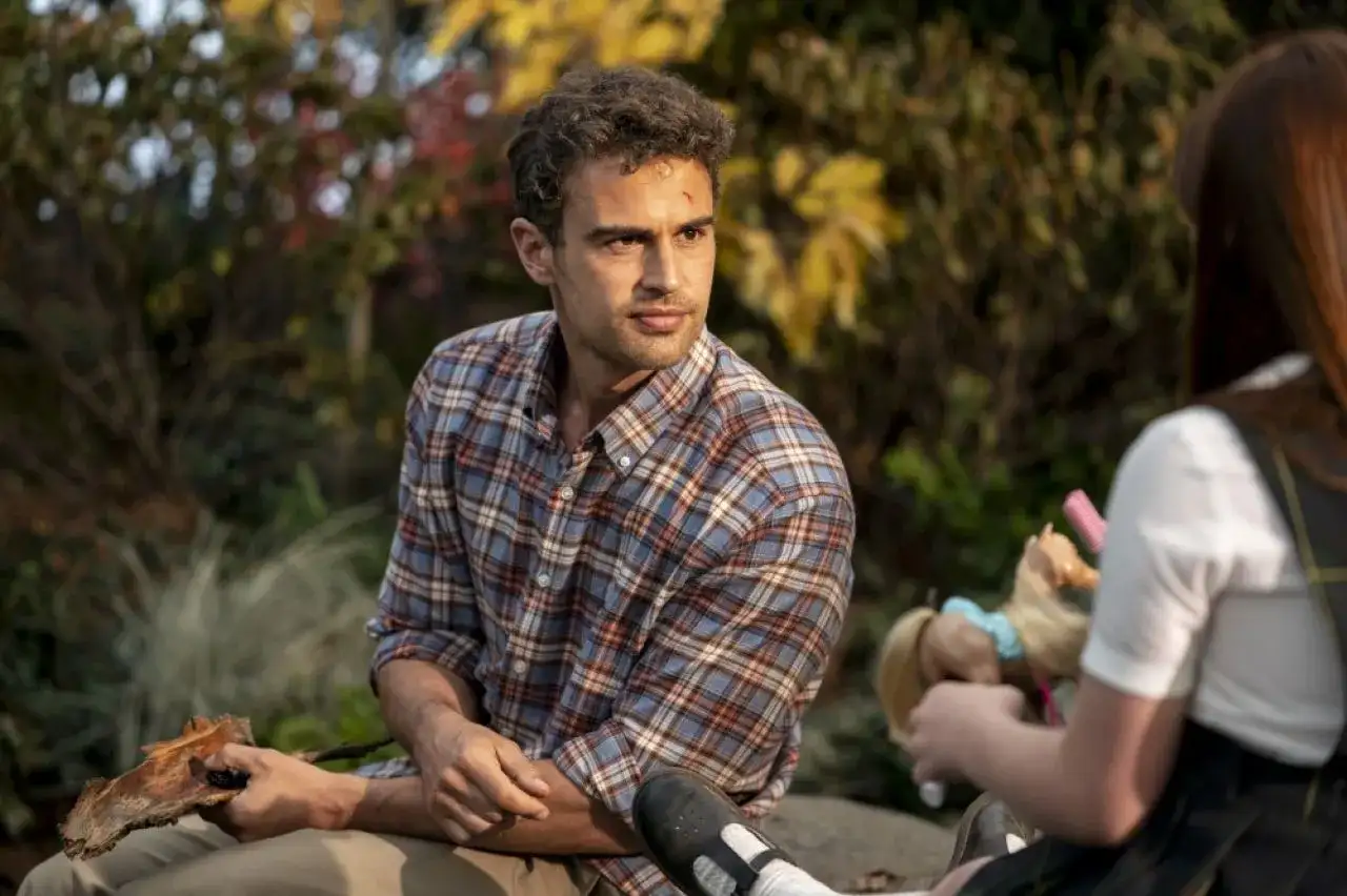 Theo James w kratowanej koszuli, znany z filmów, seriali i programów, siedzi na zewnątrz. Obok niego dziecko bawi się lalką.