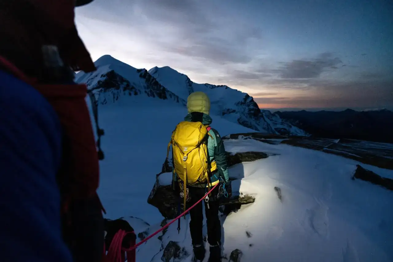 Alpinista z żółtym plecakiem lawinowym, gotowy na wyprawę w góry o wschodzie słońca. Plecak lawinowy jak działa? Zapewnia bezpieczeństwo w górach.