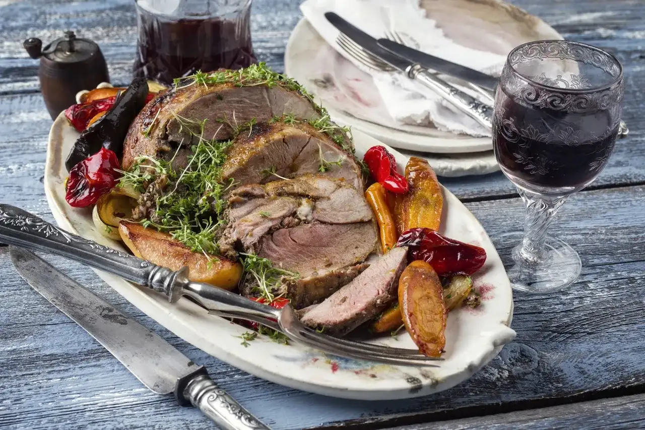 Pieczona jagnięcina z warzywami i ziołami na talerzu, obok kieliszek wina.