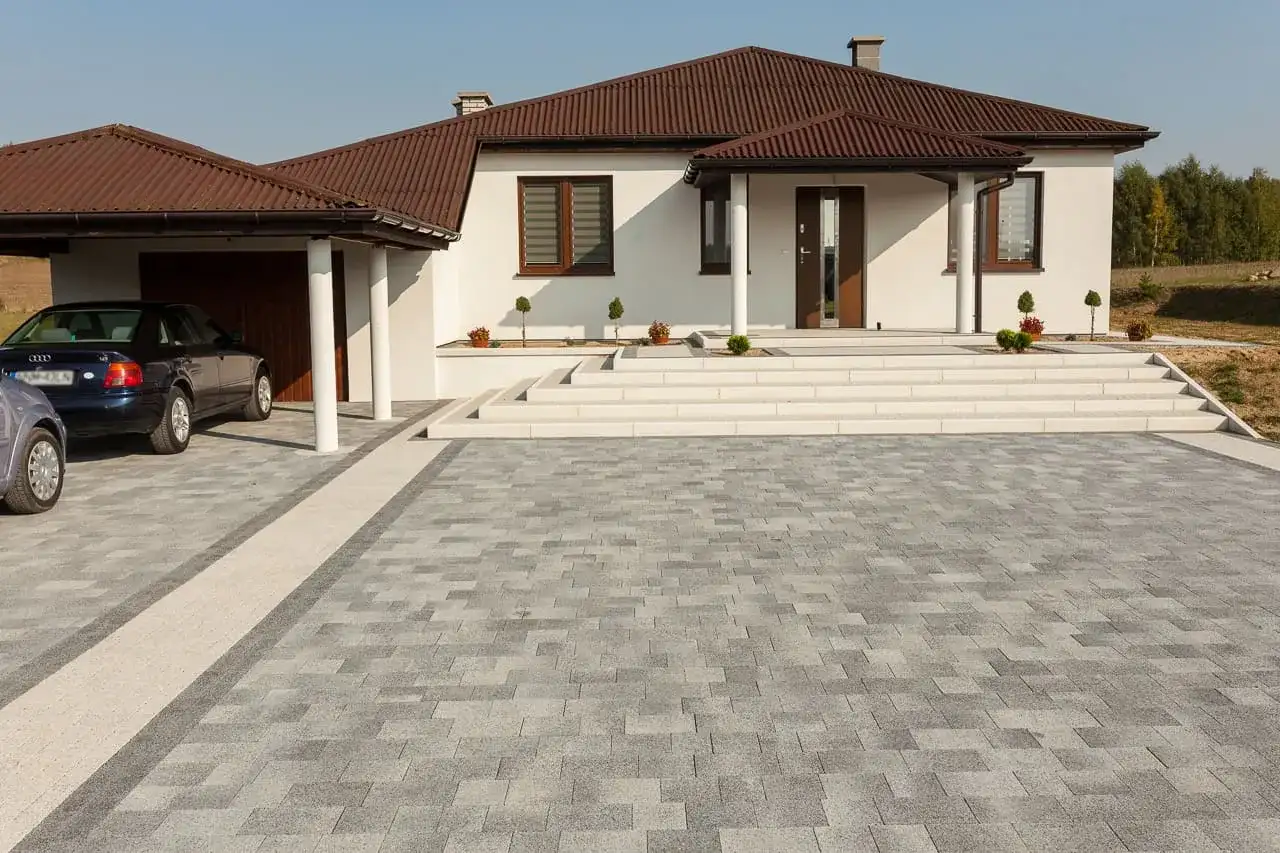 Nowoczesny podjazd z szarej kostki brukowej, idealnie komponuje się z białym domem i brązowym dachem.