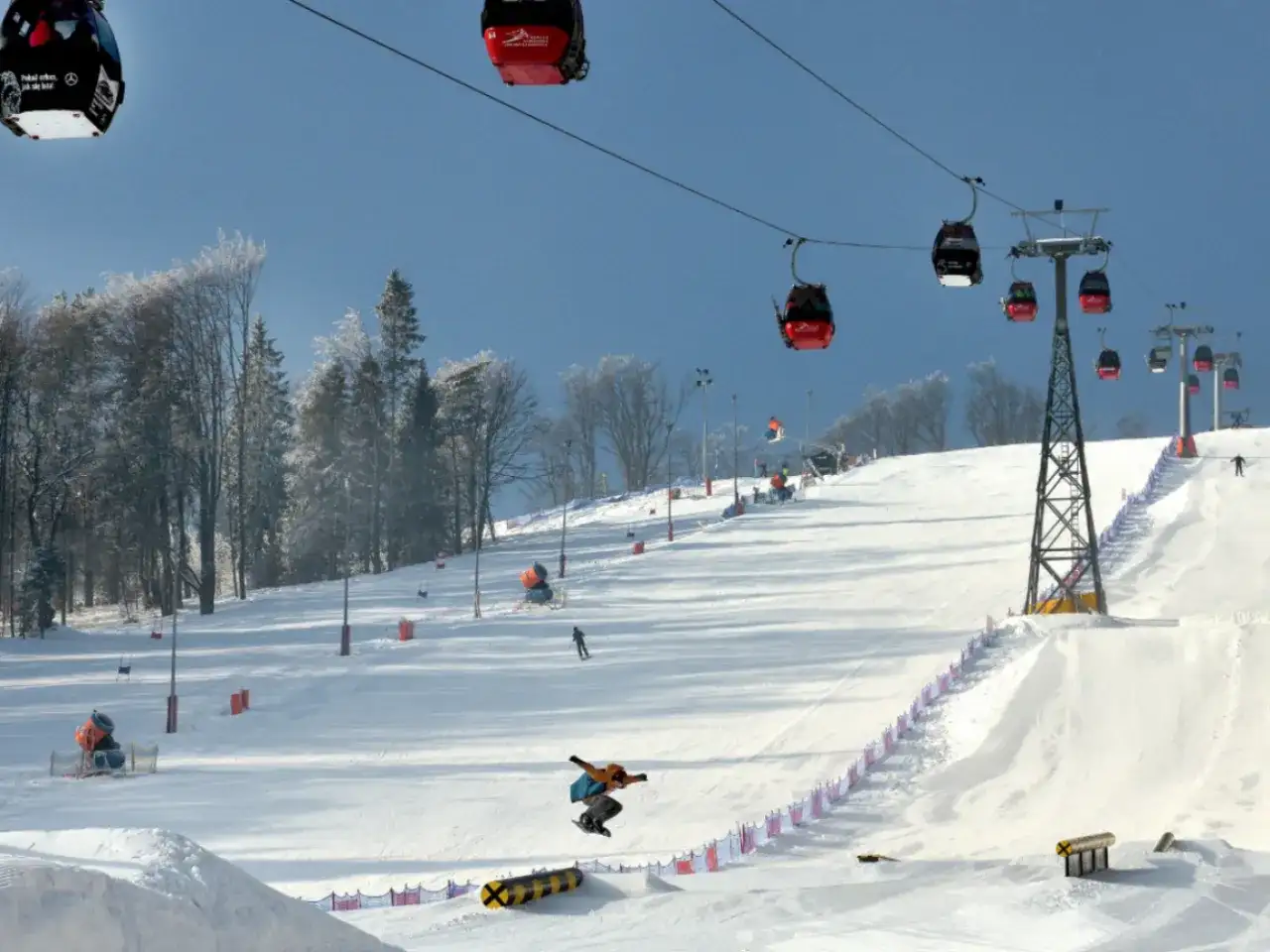 Widok na stok narciarski z kolejką linową, snowboardzistą w powietrzu i drzewami w tle.