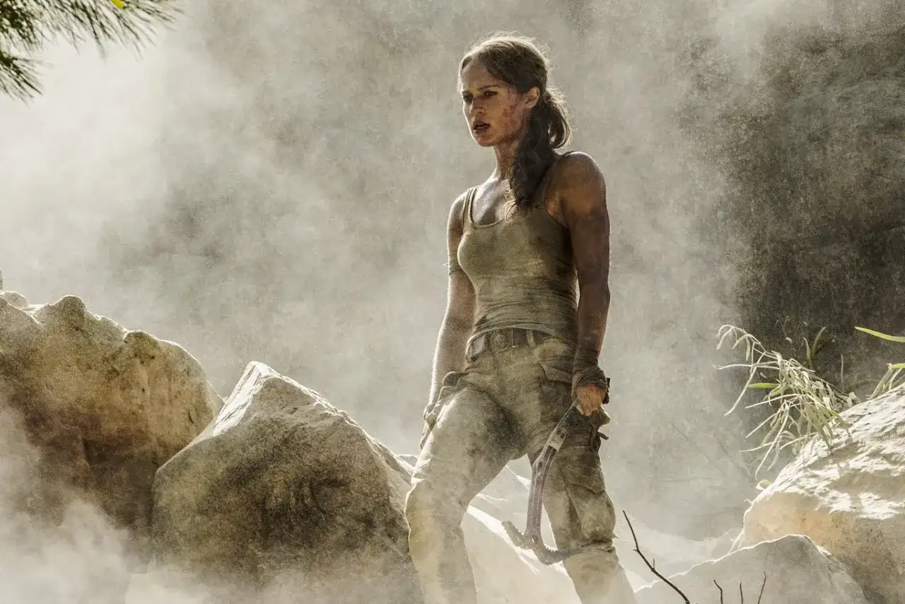 Alicia Vikander jako Lara Croft, wspinająca się po skalistym terenie, otoczona pyłem i mgłą.