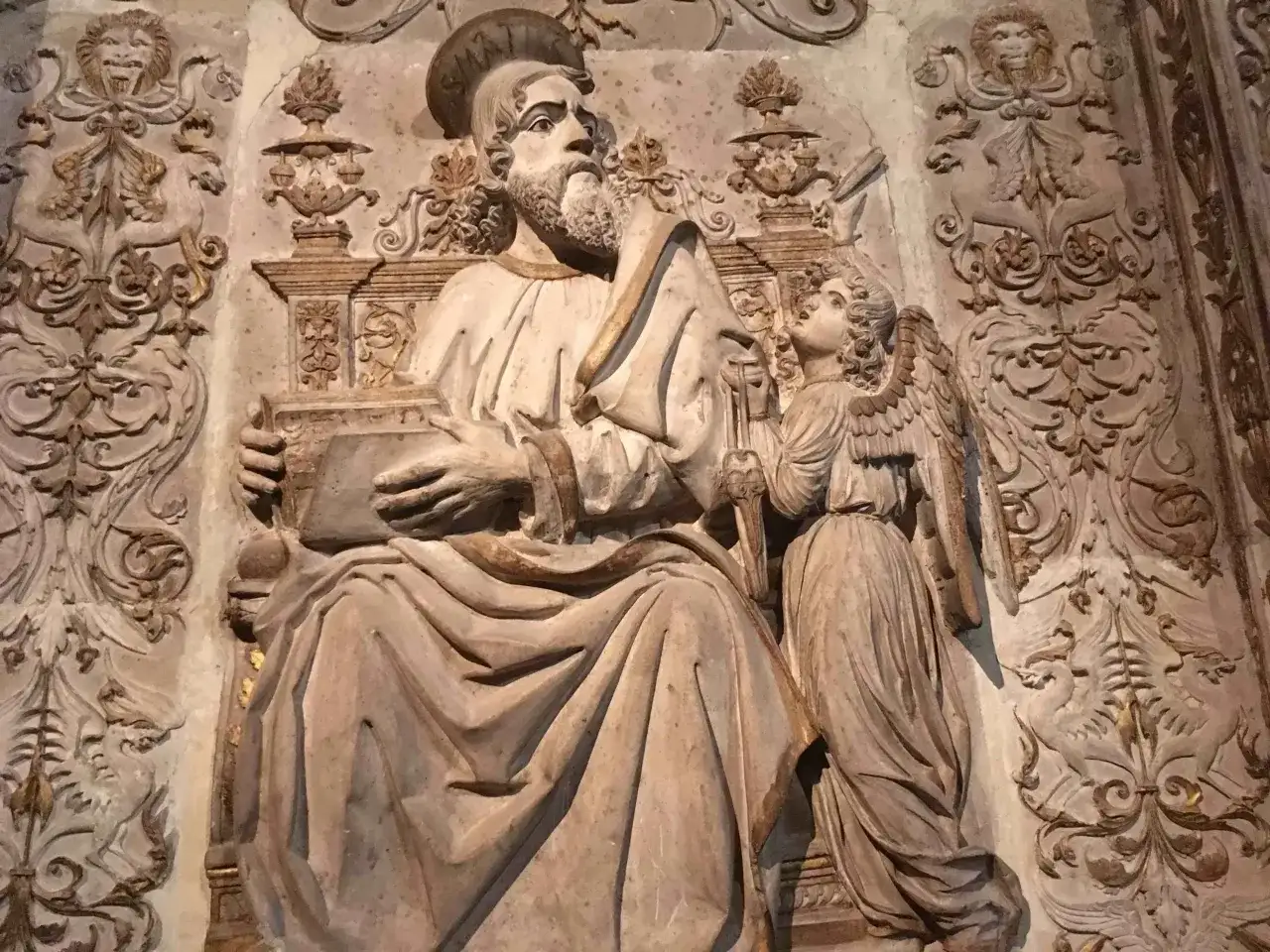 Rzeźba przedstawia św. Mateusza z aniołem, symbolizując znaczenie symboli ewangelistów. W tle widoczne ornamenty.