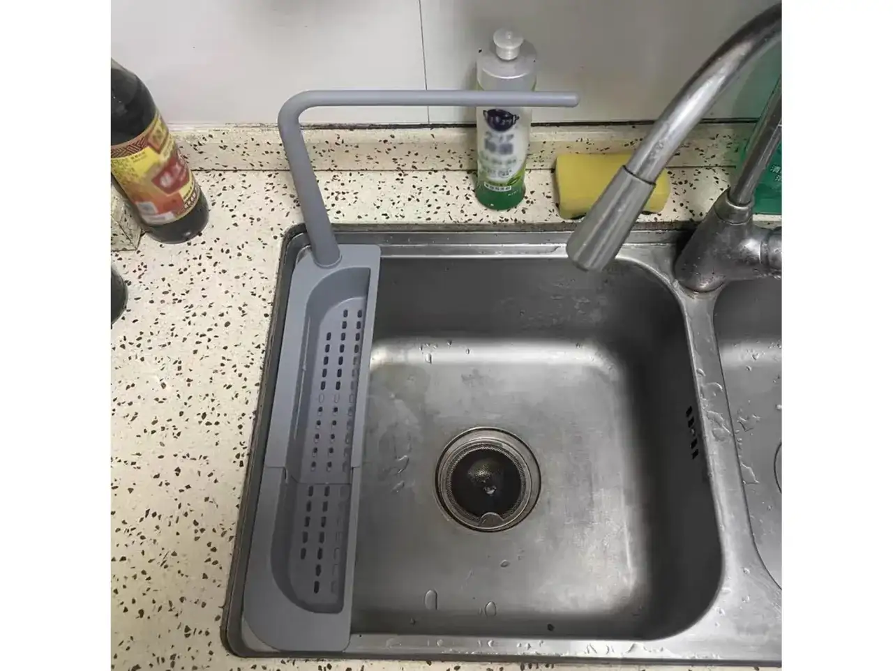 Szary uchwyt na gąbkę do zlewu, obok butelki z płynem i gąbki.