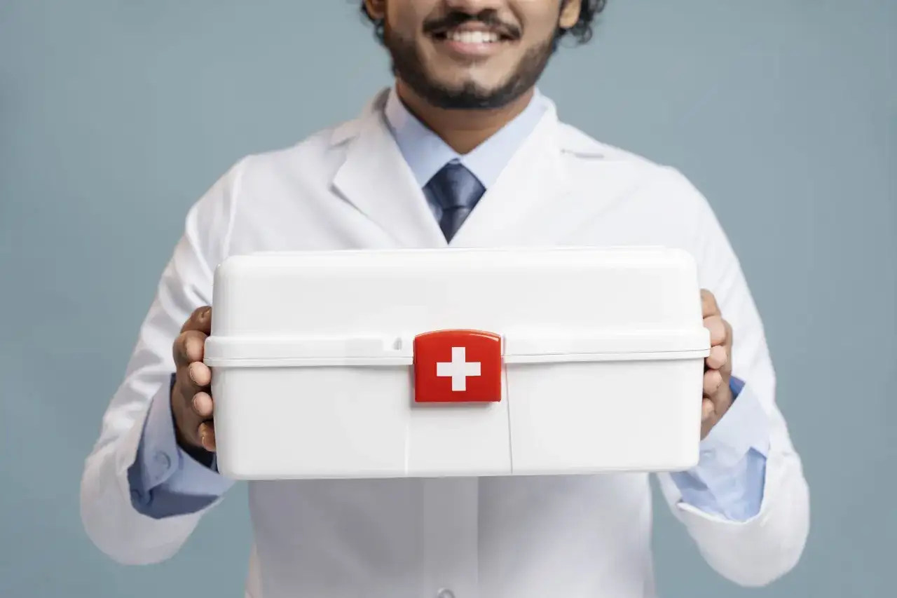 Mężczyzna w białym kitlu trzyma białą apteczkę z czerwonym krzyżem.