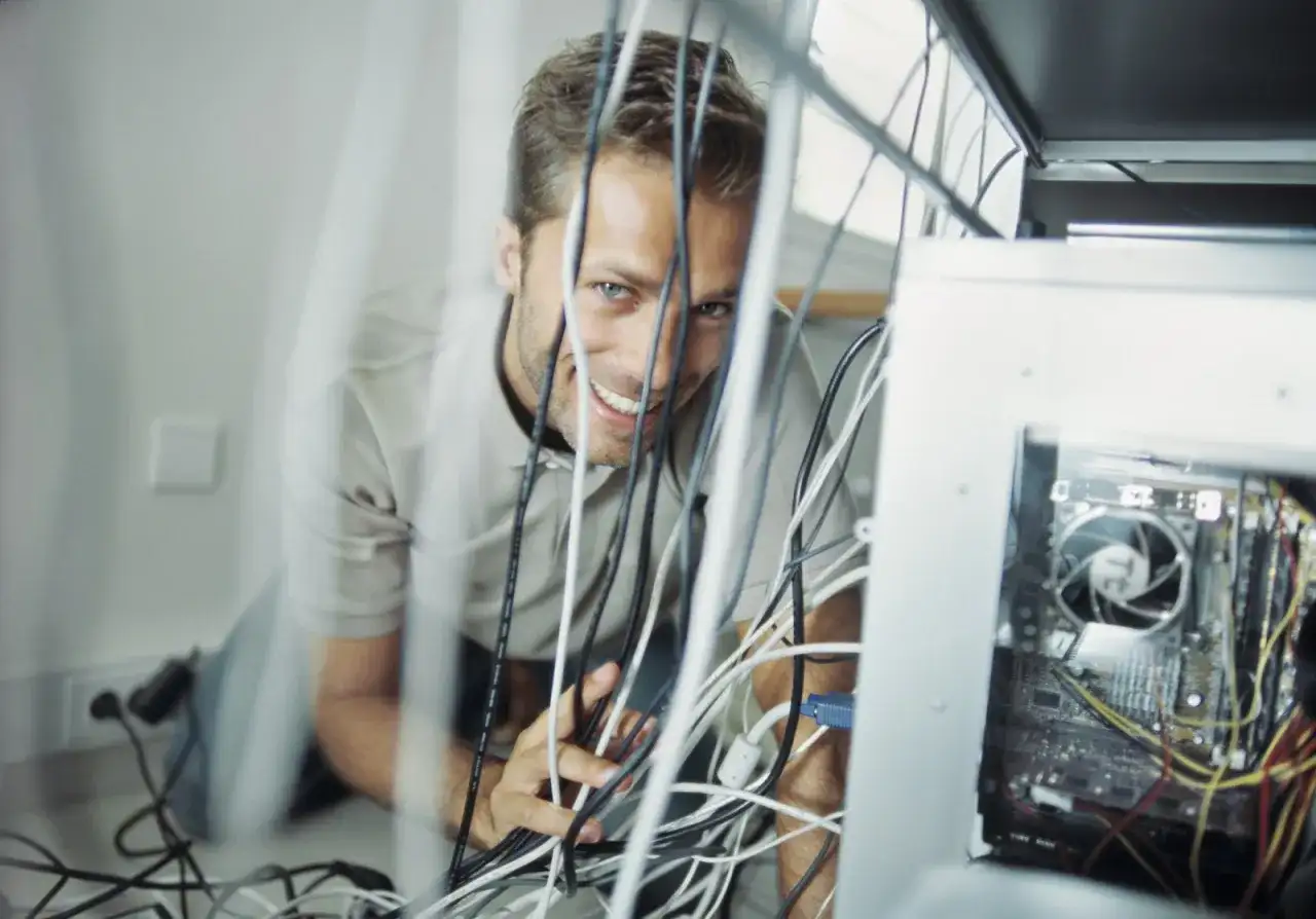 Uśmiechnięty mężczyzna grzebie w plątaninie kabli teleinformatycznych przy komputerze.