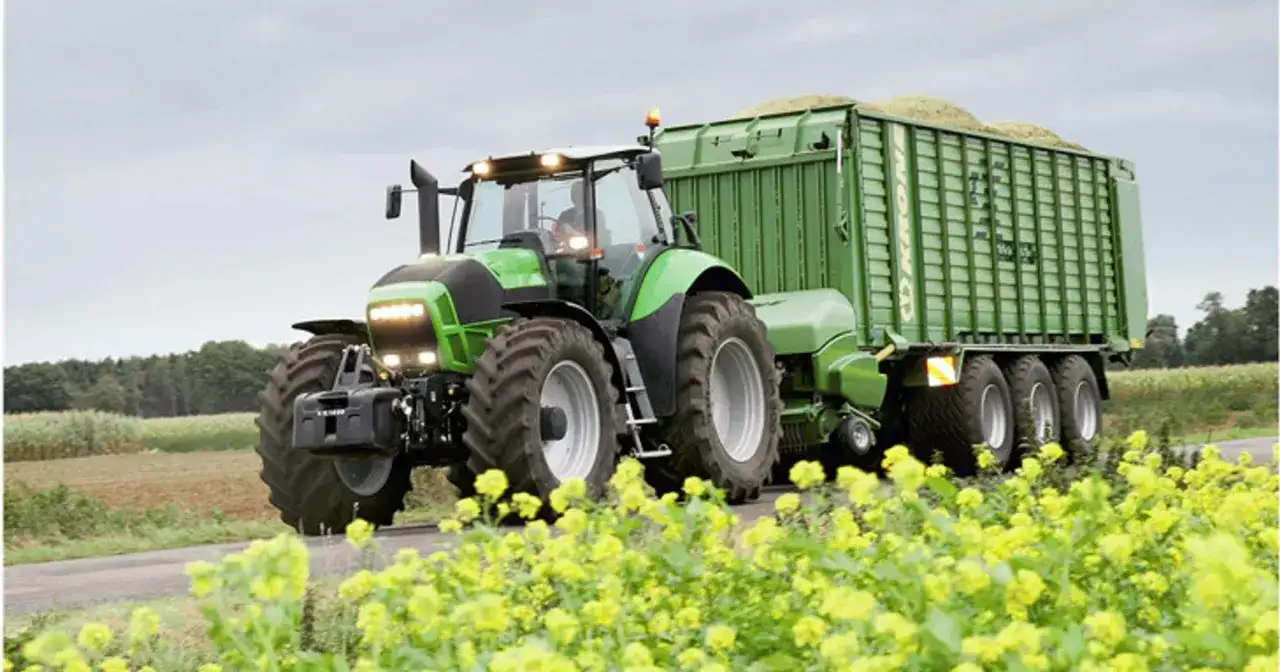 Zielony ciągnik rolniczy z przyczepą pełną zielonki na tle pola i żółtych kwiatów.