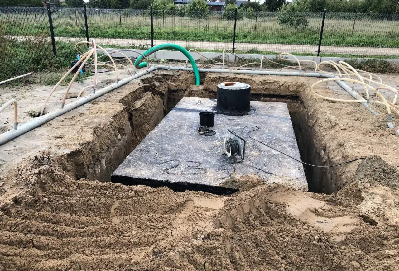 Widok na wykop pod szambo. Duża betonowa płyta, okrągły właz i liczne rury.