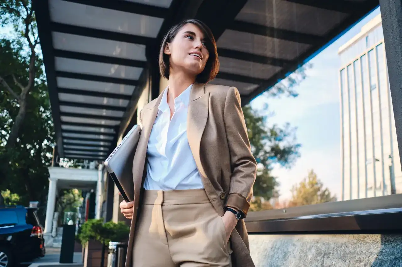 Kobieta w eleganckim stroju, symbolizującym power dressing, trzyma laptopa, gotowa na kolejne wyzwania biznesowe.