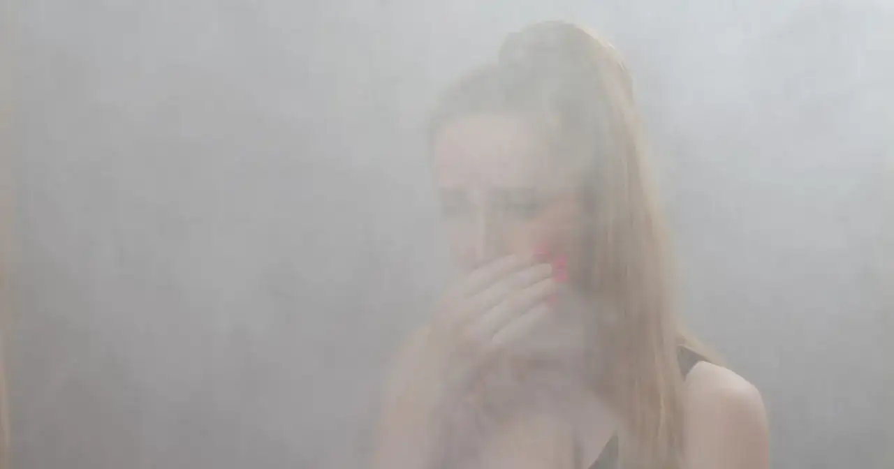 Kobieta zakrywa usta dłonią, otoczona gęstym dymem. Czy można się zatruć dymem z kominka? To pytanie nabiera tu realnego znaczenia.